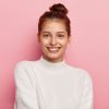 Beauty and facial expressions concept. Tender feminine woman with blue eyes, smiles pleasantly, has toothy smile, wears white comfortable sweater, looks directly at camera, isolated on pink background